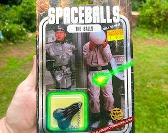 Könnte beinhalten: Eine Spaceballs Actionfigur-Karte mit dem Charakter 'The Balls', aus dessen Schritt ein grüner Laserstrahl schießt. Die Karte ist grün und schwarz mit dem Text 'Spaceballs' und 'The Balls' in weiß. Die Karte hat auch einen Aufkleber mit der Aufschrift 'Only 50 Made' und 'Kraken 4000 €'.