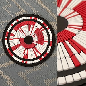 May include: A round, black coaster with a radial design in red and white. The design features a central black circle surrounded by alternating red and white segments, creating a striking visual pattern. The coaster has a black border.