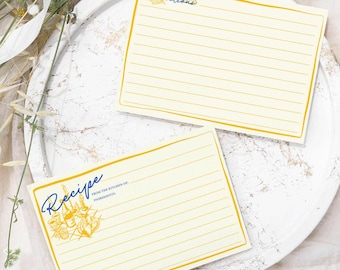 May include: Two blank recipe cards with gold trim and lined paper. The top card has the word "Directions" in blue cursive. The bottom card has the word "Recipe" in blue cursive with a gold floral design. The cards are on a white plate with a white background.