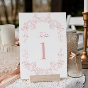Könnte beinhalten: Eine weiße Tischnummerkarte mit einem rosa Blumendesign und der Nummer 1. Die Karte wird in einem Holzhalter auf einem Tisch mit anderen dekorativen Elementen, darunter ein Kerzenhalter und eine Glasvase, angezeigt.