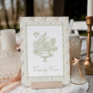 Op de afbeelding: Een witte kaart met een bloemenillustratie en de tekst "Twenty Two" op een houten standaard. De kaart heeft een delicate bloemenrand en ligt op een wit kantkleed.