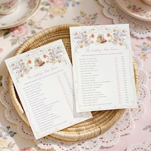 May include: Two wedding shower game cards with floral illustrations and text, resting in a woven basket. The cards are on a floral tablecloth, with teacups and saucers in the background. The text on the cards reads "The wedding shower game".