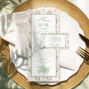 Könnte beinhalten: Ein Hochzeitsmenü und eine Tischkarte auf einem goldenen Platzteller. Das Menü zeigt ein rosa Blumenmuster mit grünen Akzenten und das Wort "Menü" oben. Die Tischkarte trägt den Namen "Charlotte".