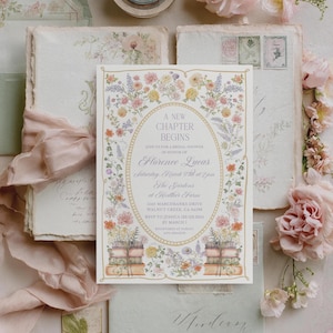 Pode incluir: Um conjunto de convites de casamento com um design floral. O cartão de convite tem o texto "A New Chapter Begins" dentro de uma borda floral. O conjunto inclui envelopes, papelaria e fita rosa, com flores espalhadas.