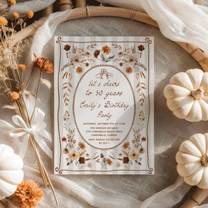 May include: A rectangular invitation card with a floral design and the text "let's cheers to 30 years Emily's Birthday Party". The card has a striped background and is surrounded by dried flowers and small white pumpkins.