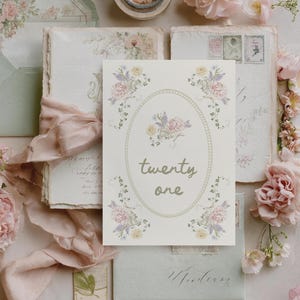Pode incluir: Uma composição elegante com elementos de papelaria e florais. Um cartão com o texto "twenty one" é rodeado por desenhos florais. Envelopes, papelaria e flores em tons suaves de rosa e verde são dispostos sobre uma superfície neutra.