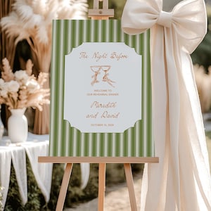May include: A wooden easel holds a green and white striped sign with the text "The Night Before" and "Welcome to Our Rehearsal Dinner". The sign also includes the names "Meredith and David" and the date "October 10, 2024". A large cream-colored bow is attached to the easel.