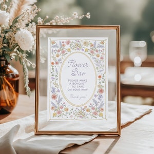 May include: A framed sign with the text "Flower Bar" and floral illustrations. The sign is in a gold frame and features an oval design with the text "Please make a bouquet to take on your way! Thank you!" A vase of flowers is in the background.