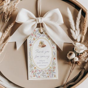 May include: A white satin bow tied around a string attached to a white card with a floral border. The card has a purple oval with the text "Thank you for coming" and "Emily's Baby Shower 03/03/2023".