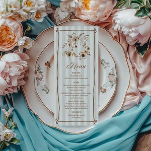 May include: A wedding menu card with a floral design, placed on a white plate with gold trim and floral accents. The menu card has a striped background and features the word "Menu" at the top. The scene is surrounded by pink and white flowers and blue fabric.
