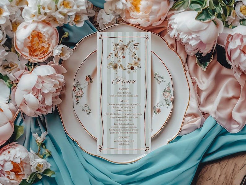 May include: A wedding menu card with a floral design, placed on a white plate with gold trim and floral accents. The menu card has a striped background and features the word "Menu" at the top. The scene is surrounded by pink and white flowers and blue fabric.