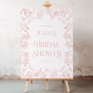 May include: A rectangular sign on a wooden easel featuring a white background with a dusty pink botanical border. The text reads: WELCOME TO JULIA'S BRIDAL SHOWER MARCH 6, 20XX. The floral illustrations frame the central text in a symmetrical pattern.