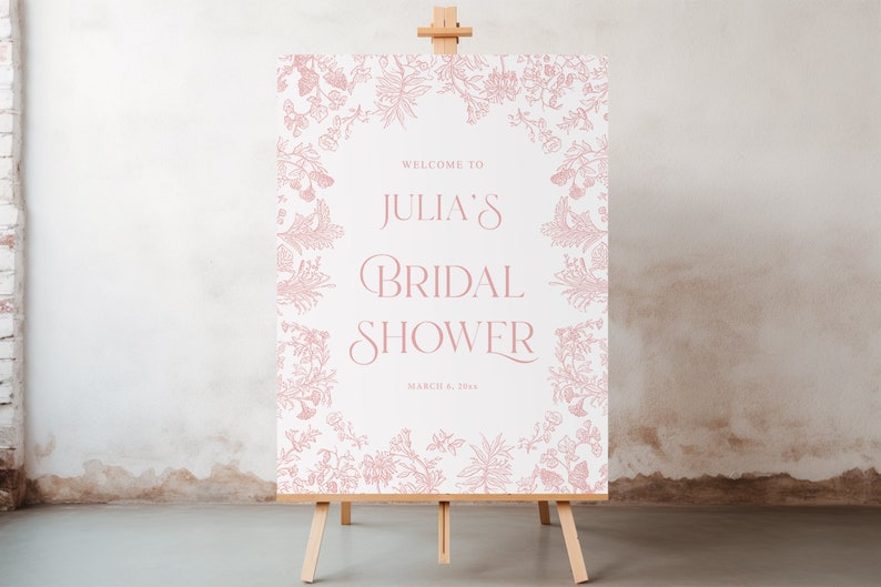 May include: A rectangular sign on a wooden easel featuring a white background with a dusty pink botanical border. The text reads: WELCOME TO JULIA'S BRIDAL SHOWER MARCH 6, 20XX. The floral illustrations frame the central text in a symmetrical pattern.