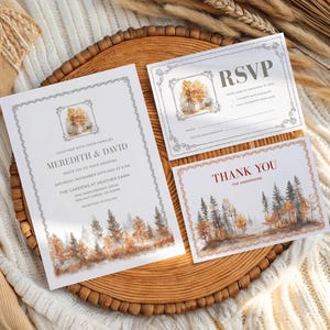 May include: A set of wedding stationery featuring a watercolor forest design. The invitation, RSVP, and thank you cards have a decorative border and include the names Meredith & David. The cards are on a wooden tray.
