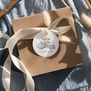 May include: A brown gift box with a white ribbon and a round thank you tag. The tag features a blue and white teapot with a tiered cake stand and the text "Thank You".