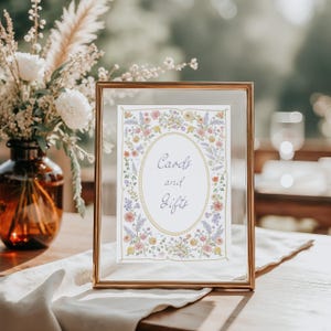 Pu&ograve; includere: Un cartello dorato con la scritta "Cards and Gifts" in un ovale floreale. La cornice &egrave; dorata e il cartello &egrave; su un tavolo di legno con un panno bianco. Un vaso di fiori &egrave; sullo sfondo, per un evento speciale.
