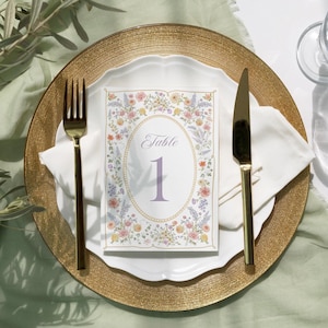 Op de afbeelding: Een elegante tafelsetting met een gouden onderbord, een wit bord en een bloemrijke tafelnumerkaart. De kaart toont een ovale lijst met bloemen, het woord "Table" en het nummer "1". Gouden bestek en een witte servet maken de opstelling compleet.