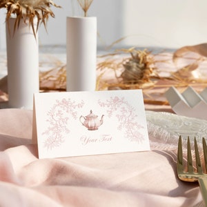 May include: A white place card with a pink teapot illustration and the text "Your Text". The place card is on a pink tablecloth with a gold fork and a white plate.