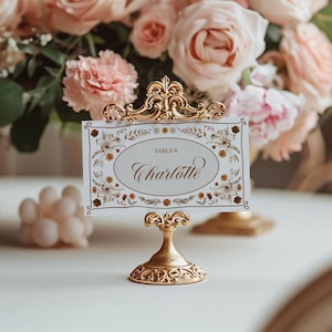 May include: A gold-colored table card holder with an ornate design, holding a card that reads "Table 6 Charlotte." The card has a floral border. Soft pink roses and other flowers are in the background.