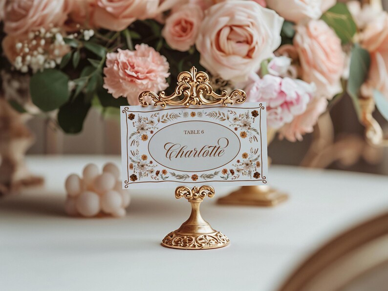 May include: A gold-colored table card holder with an ornate design, holding a card that reads "Table 6 Charlotte." The card has a floral border. Soft pink roses and other flowers are in the background.