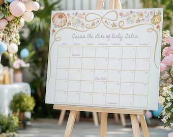 May include: A baby shower calendar on a wooden easel. The calendar is white with gold lettering and floral accents. The text reads "Guess the date of baby Julia." The calendar grid is visible, with the word "due date" written on it.