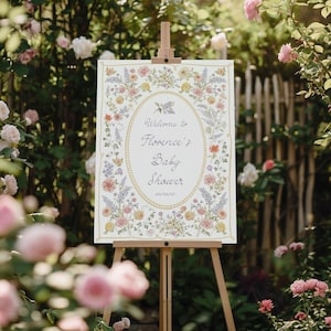 Pu&ograve; includere: Un cartello per baby shower su un cavalletto di legno, circondato da fiori. Il cartello ha un bordo floreale e un centro ovale con il testo "Welcome to Florence's Baby Shower" e la data 04/04/23.