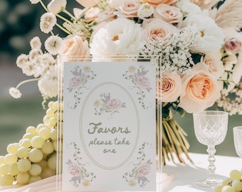 Könnte beinhalten: Ein klarer Acrylschild mit dem Text "Favors please take one" in einer dekorativen Schriftart, umgeben von floralen Illustrationen. Ein Strauß aus weißen und pfirsichfarbenen Rosen und anderen Blumen befindet sich im Hintergrund, zusammen mit einer Traube grüner Trauben und Kristallgläsern.