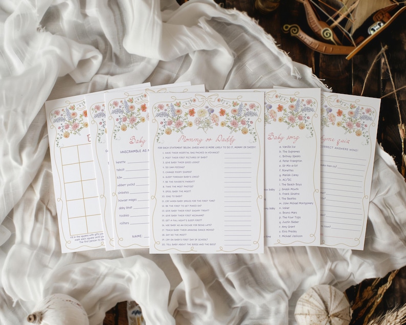 Pu&ograve; includere: Un set di biglietti per giochi per baby shower con decorazioni floreali. Le carte presentano titoli come "Baby Unscramble" e "Mommy or Daddy". Il bordo floreale include fiori rosa, arancioni e viola. Le carte sono disposte su un tessuto bianco.