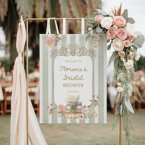 May include: A sign for a bridal shower with the text "Welcome to Florence's Bridal Shower 10/10/202x" in elegant script. The sign has a striped background with floral and book illustrations, and is decorated with roses and greenery.