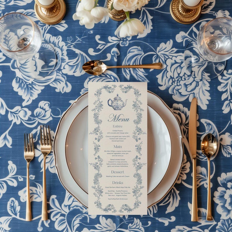May include: A white menu card with a blue teapot illustration and floral border. The menu lists "Entree", "Main", "Dessert", and "Drinks" with descriptions of each item. The card is placed on a white plate with a gold rim, which is on a blue and white floral tablecloth.