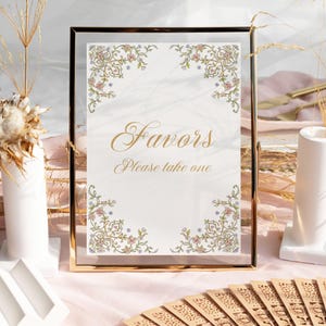 Op de afbeelding: Een gouden lijst met een bloemenontwerp en de tekst "Favors Please take one". Het bord staat op een tafel met een witte vaas en een waaier.