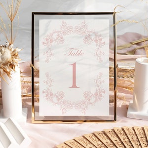 May include: A rectangular table number sign featuring the text Table 1 in a soft pink font, centered within a delicate pink floral wreath illustration. The sign is encased in a thin, metallic gold frame with a clear glass border.