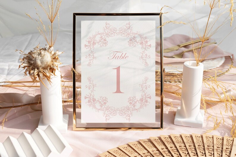 May include: A rectangular table number sign featuring the text Table 1 in a soft pink font, centered within a delicate pink floral wreath illustration. The sign is encased in a thin, metallic gold frame with a clear glass border.