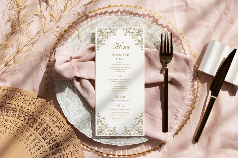 May include: A vertical white menu card with gold and floral border details, featuring the text Menu, Alternate Drop, Entr&eacute;e, Main, Dessert, and Drinks. It is placed on a pink napkin, a clear glass charger plate, a black fork, and a black knife.