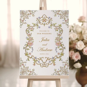 May include: A white wedding sign with a gold ornate frame and floral accents. The sign reads "Welcome to our wedding Julia and Andrew March 6, 2024".