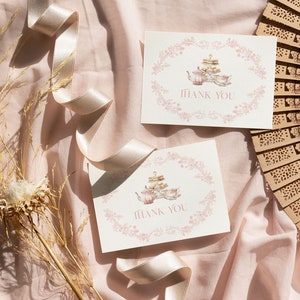 May include: Two white thank you cards with pink floral designs and the text "Thank You". The cards feature a watercolor illustration of a teacup and teapot. The cards are on a pink fabric background with a wooden fan and a ribbon.