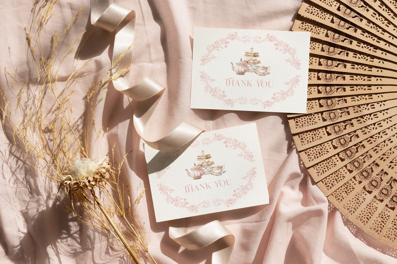 May include: Two white thank you cards with pink floral designs and the text "Thank You". The cards feature a watercolor illustration of a teacup and teapot. The cards are on a pink fabric background with a wooden fan and a ribbon.