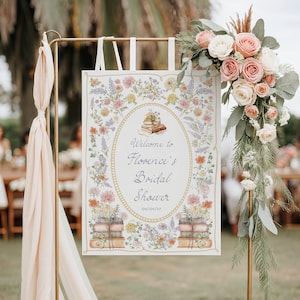 May include: A rectangular sign hanging from a gold metal stand, featuring a floral border with watercolor illustrations of books and the text Welcome to Florence's Bridal Shower 04/04/27. The sign is decorated with a large bouquet of pink and white roses.