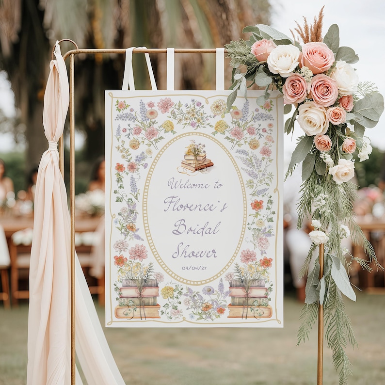May include: A rectangular sign hanging from a gold metal stand, featuring a floral border with watercolor illustrations of books and the text Welcome to Florence's Bridal Shower 04/04/27. The sign is decorated with a large bouquet of pink and white roses.