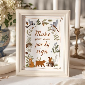 Op de afbeelding: Een witte rechthoekige fotolijst met een bordje met de tekst "Make your own party sign" in bruin handschrift. Het bordje bevat illustraties van vogels, eikels, dennentakken, een vos, een beer, een eekhoorn, een egel en een konijn.