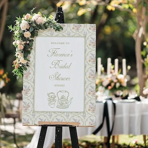 K&ouml;nnte beinhalten: Ein dekoratives Schild auf einer Staffelei, das die G&auml;ste zu &bdquo;Florence's Bridal Shower&ldquo; am 10.10.2027 willkommen hei&szlig;t. Das Schild hat einen floralen Rahmen und Illustrationen einer Teekanne und einer Tasse. Ein Blumenarrangement schm&uuml;ckt die linke obere Ecke.