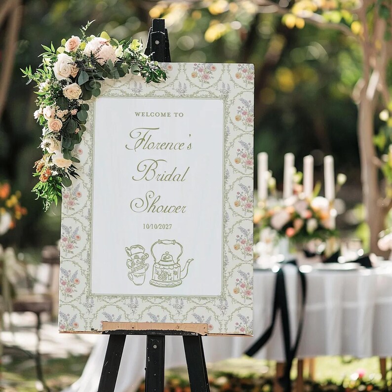 K&ouml;nnte beinhalten: Ein dekoratives Schild auf einer Staffelei, das die G&auml;ste zu &bdquo;Florence's Bridal Shower&ldquo; am 10.10.2027 willkommen hei&szlig;t. Das Schild hat einen floralen Rahmen und Illustrationen einer Teekanne und einer Tasse. Ein Blumenarrangement schm&uuml;ckt die linke obere Ecke.