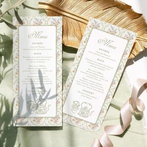 May include: Two wedding menu cards with a floral border and a delicate design. The menu features a three-course meal with chilled prawns, chorizo and green pea risotto, and rib fillet. The dessert is wedding cake served with cream.