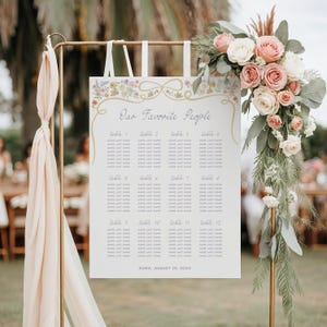 May include: A white wedding seating chart with a floral border and gold ribbon accents. The chart lists table numbers and guest names. The text reads "Our Favorite People" and "ROMA, AUGUST 15, 20XX".