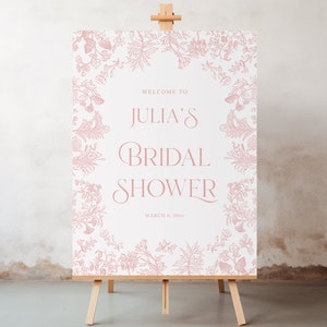 May include: A white bridal shower sign with a pink floral border. The sign reads "Welcome to Julia's Bridal Shower, March 8, 2024".