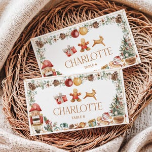 May include: Two rectangular white place cards featuring a watercolour Christmas border with pinecones, ornaments, a gingerbread person, a reindeer, a nutcracker, and a tree. The text reads CHARLOTTE and TABLE 6 in gold lettering.