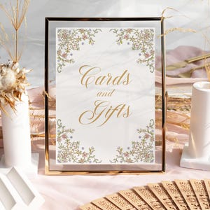 Op de afbeelding: Een gouden lijst met een witte achtergrond en een bloemige rand. Het bord heeft de tekst "Kaarten en Geschenken" in gouden letters.