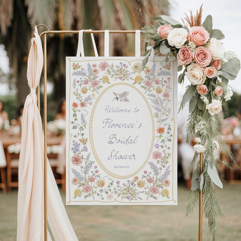 May include: A sign for a bridal shower, featuring the text "Welcome to Florence's Bridal Shower" within an oval frame. The sign is decorated with floral patterns and is supported by a gold metal stand. A floral arrangement of roses and greenery adorns the top.