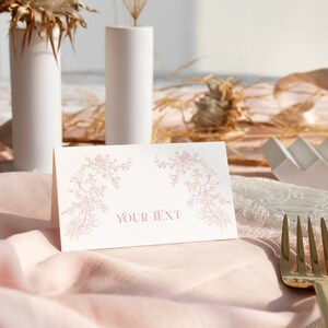 May include: A folded white paper place card featuring a delicate pink floral vine illustration on the left and right sides, with the centered text "YOUR TEXT" printed in a matching pink serif font, positioned on a soft pink fabric napkin.