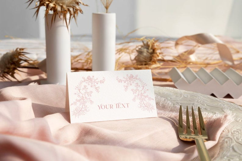May include: A folded white paper place card featuring a delicate pink floral vine illustration on the left and right sides, with the centered text "YOUR TEXT" printed in a matching pink serif font, positioned on a soft pink fabric napkin.